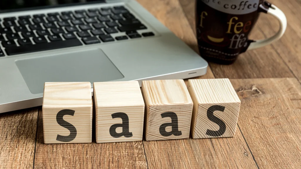 Wooden blocks spelling "SaaS" placed on a wooden desk next to a laptop and coffee cup.