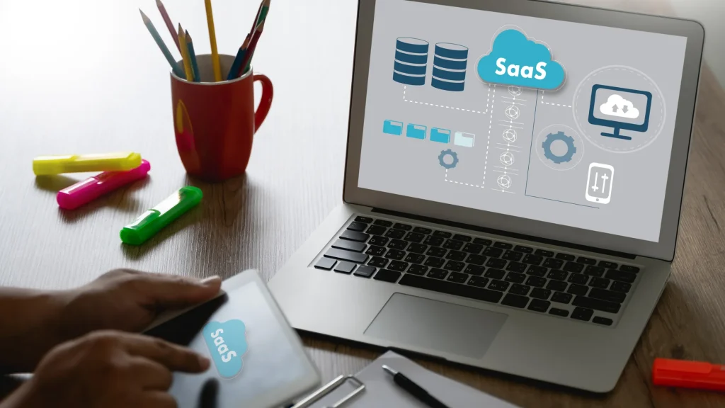 A person using a tablet with SaaS cloud service graphics on a laptop screen in a workspace.